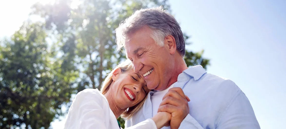 A smiling older couple dancing outdoors under sunlit trees
